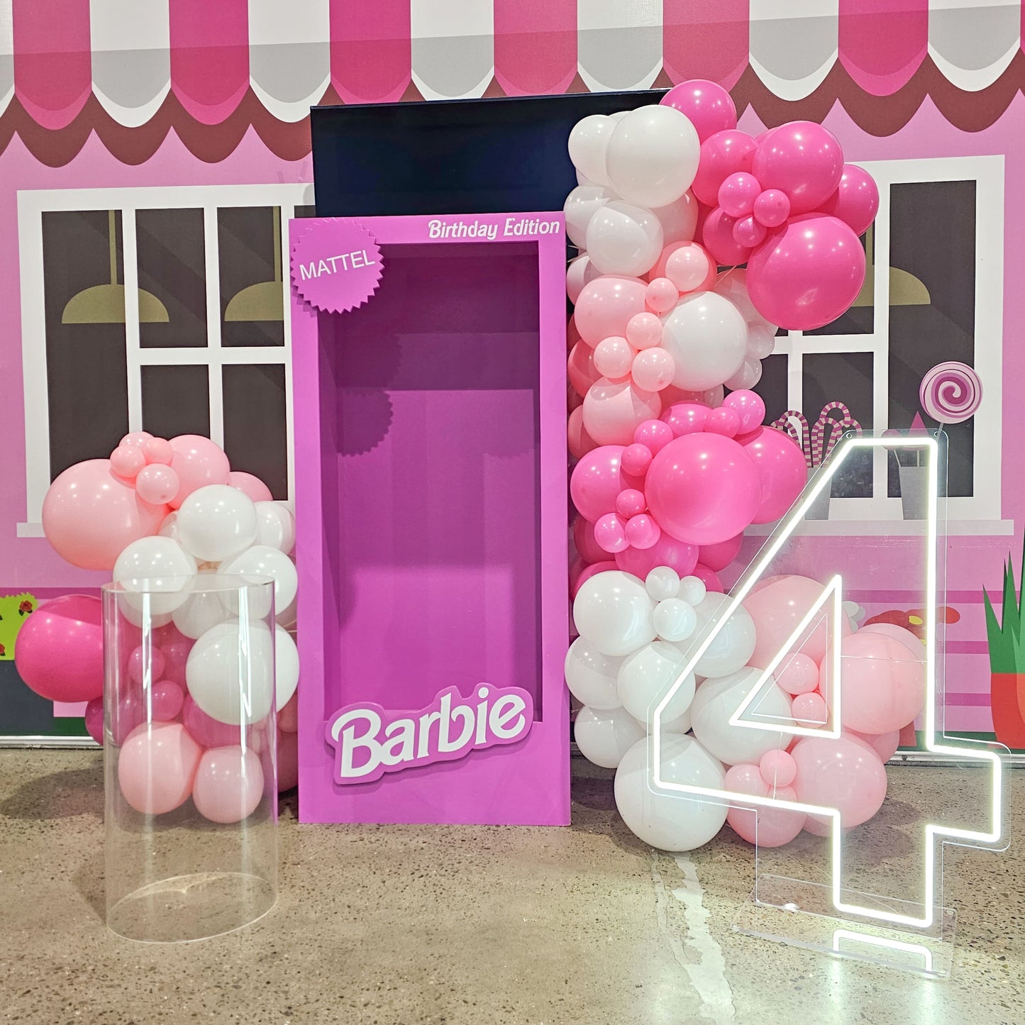 Child sized doll photo booth in hot pink with large neon number, balloon garland decoration and clear acrylic plinth for a play centre birthday party.