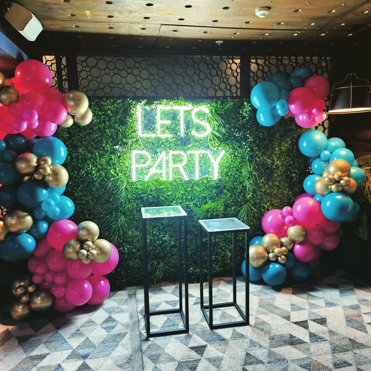 Lets party neon sign on a green wall backdrop with organic balloon garland decor and 2 black steel cake plinths for a birthday party photo opportunity