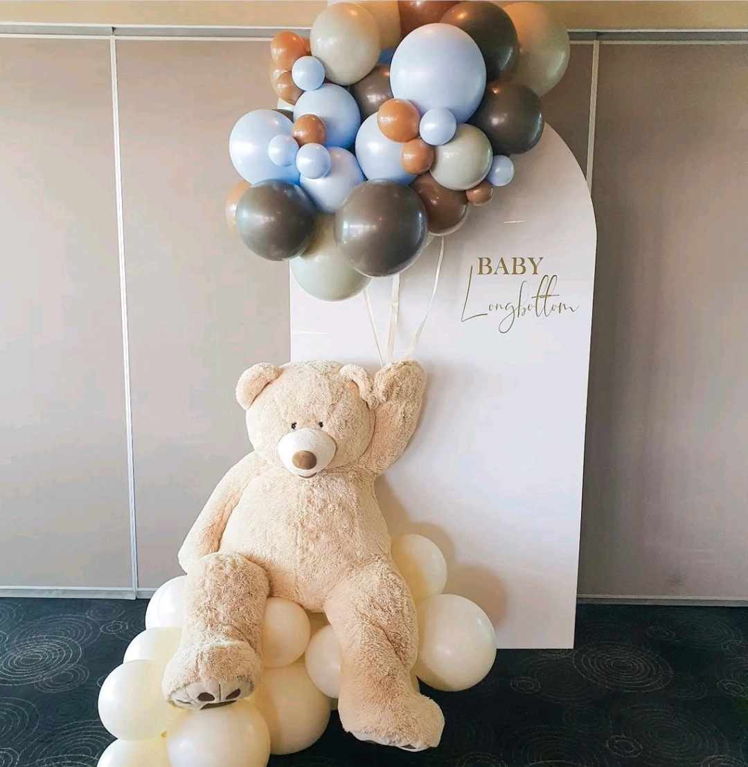 Lifesize teddy bear sitting on a balloon cloud holding a bouquet of balloons, featuring our white arch backdrop for a We can bearly wait baby shower decoration