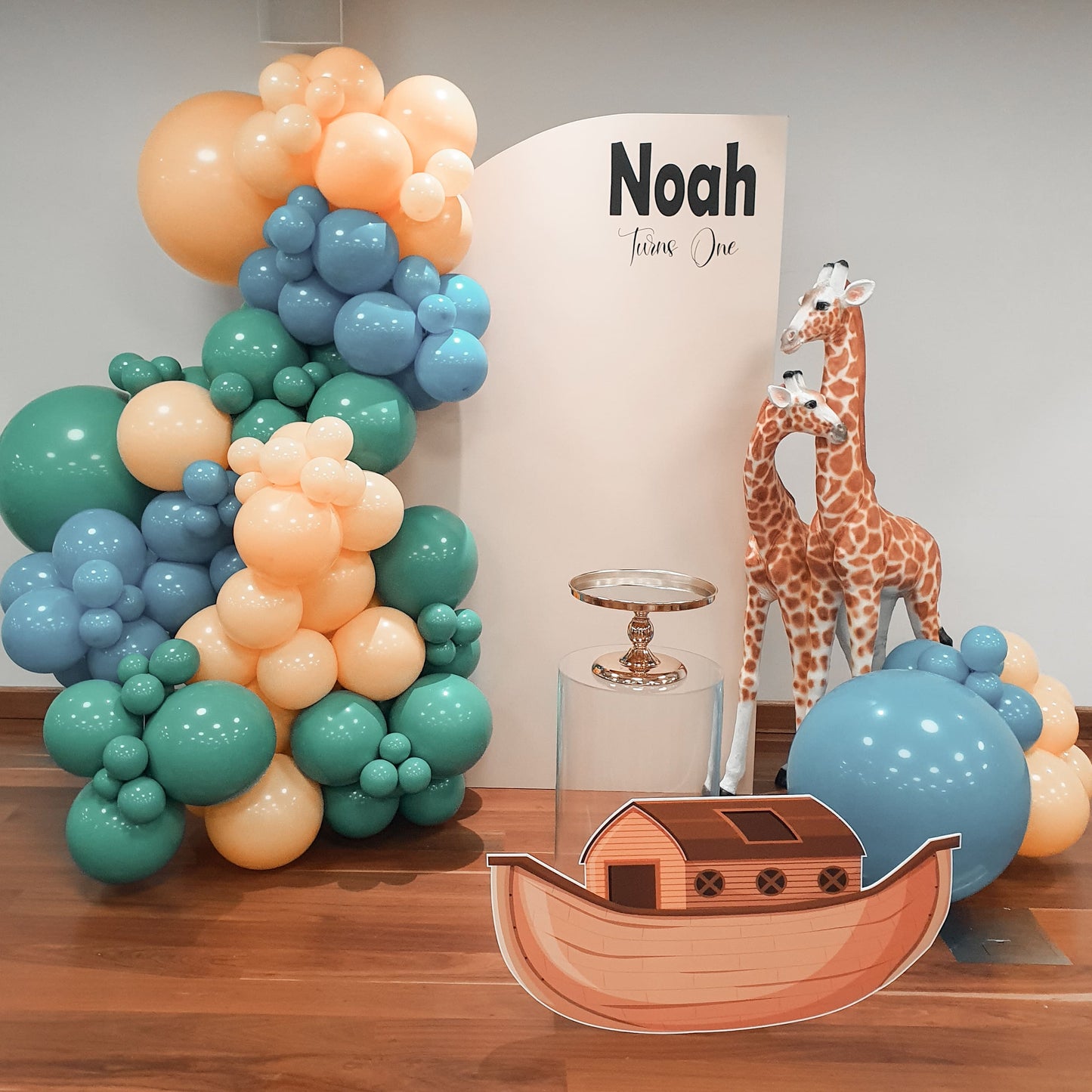 Sail arch backdrop, noahs ark cutout, clear acrylic plinth, gold cake stand and safari animals with balloon garland for a Noahs ark themed first birthday in Sydney.