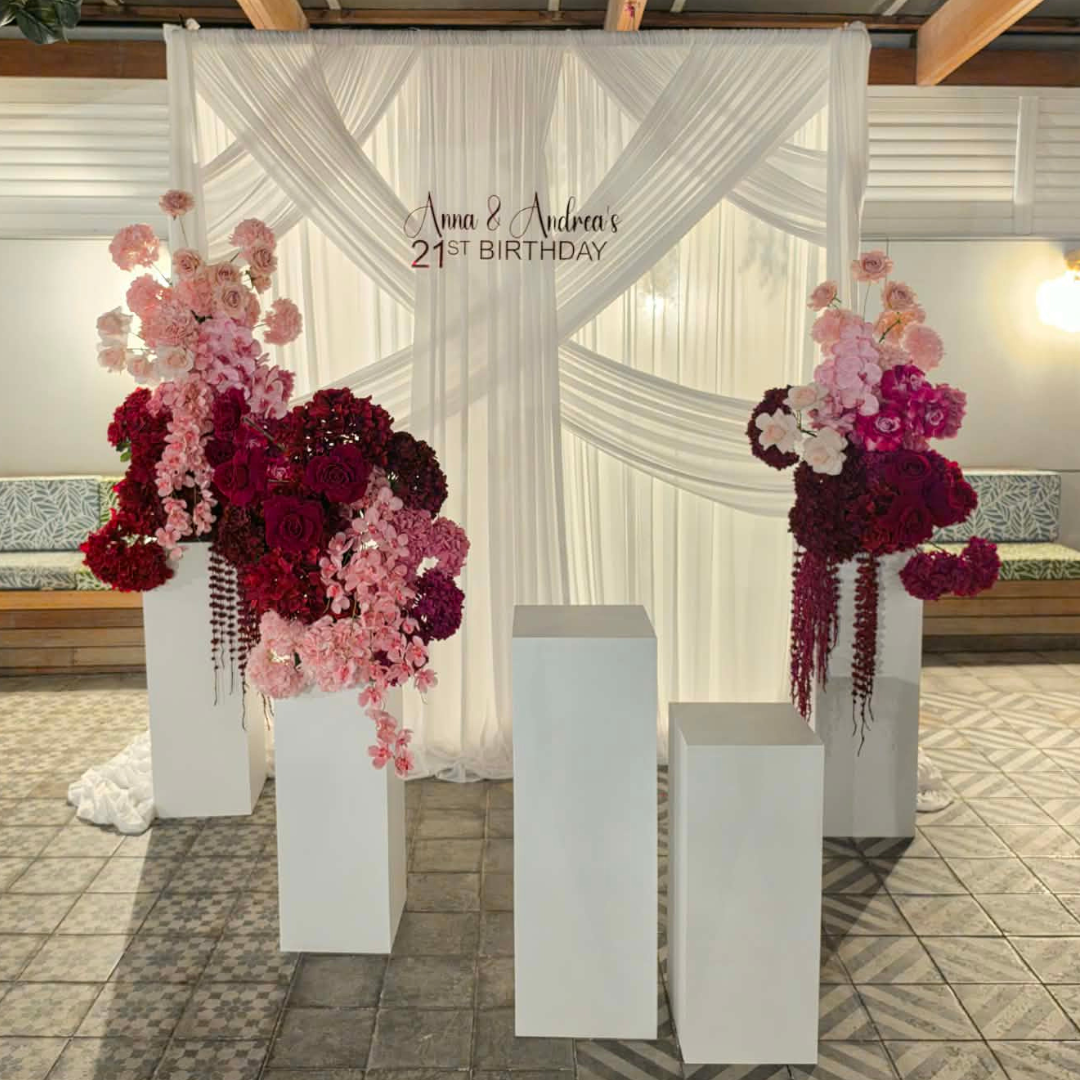 Decorative floral arrangements on white plinths with 'Anna & Andrea's 21st Birthday' text in the background.