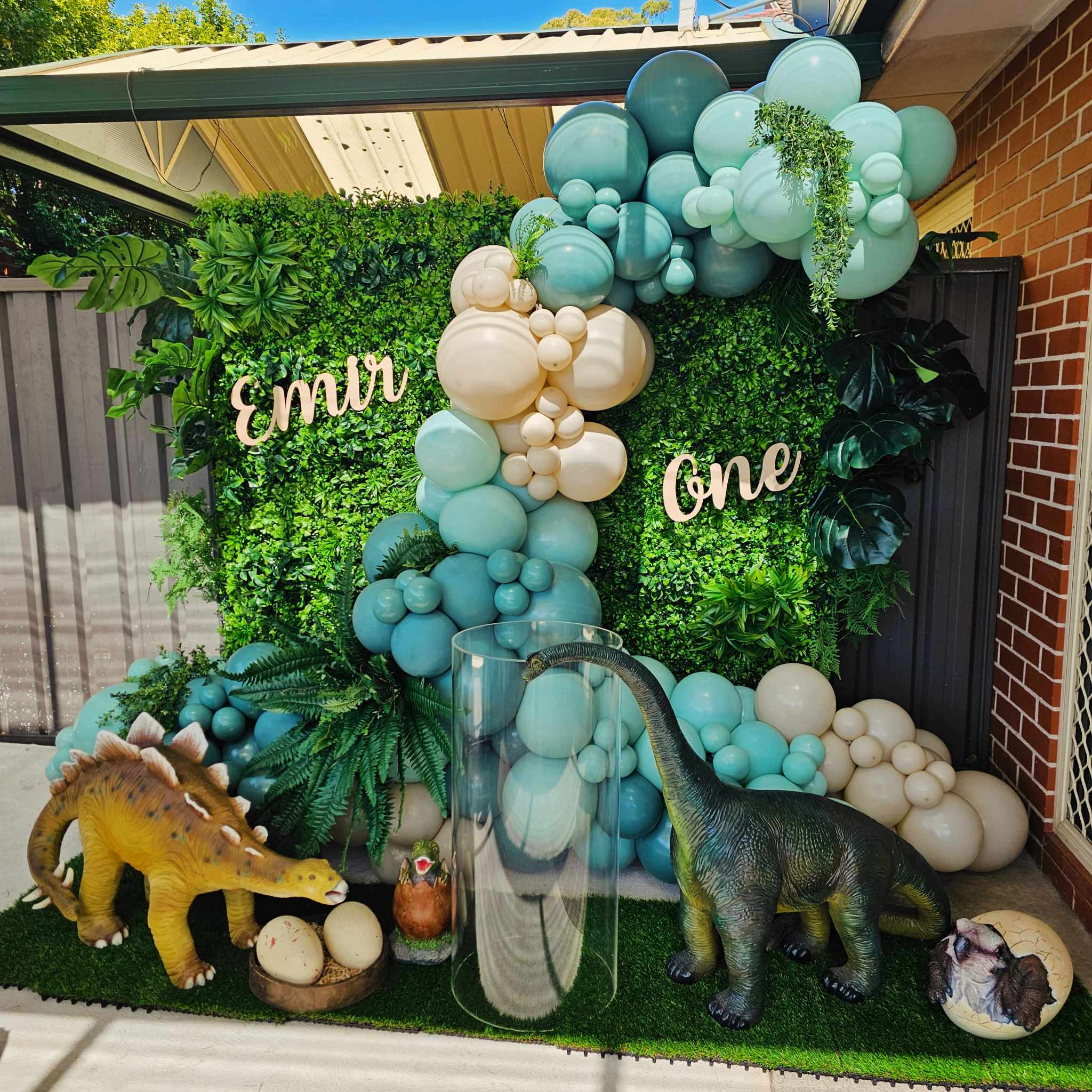 Green wall textured backdrop for first birthday party with dinosaur props, clear acylic plinth to display the cake and organic balloon garland