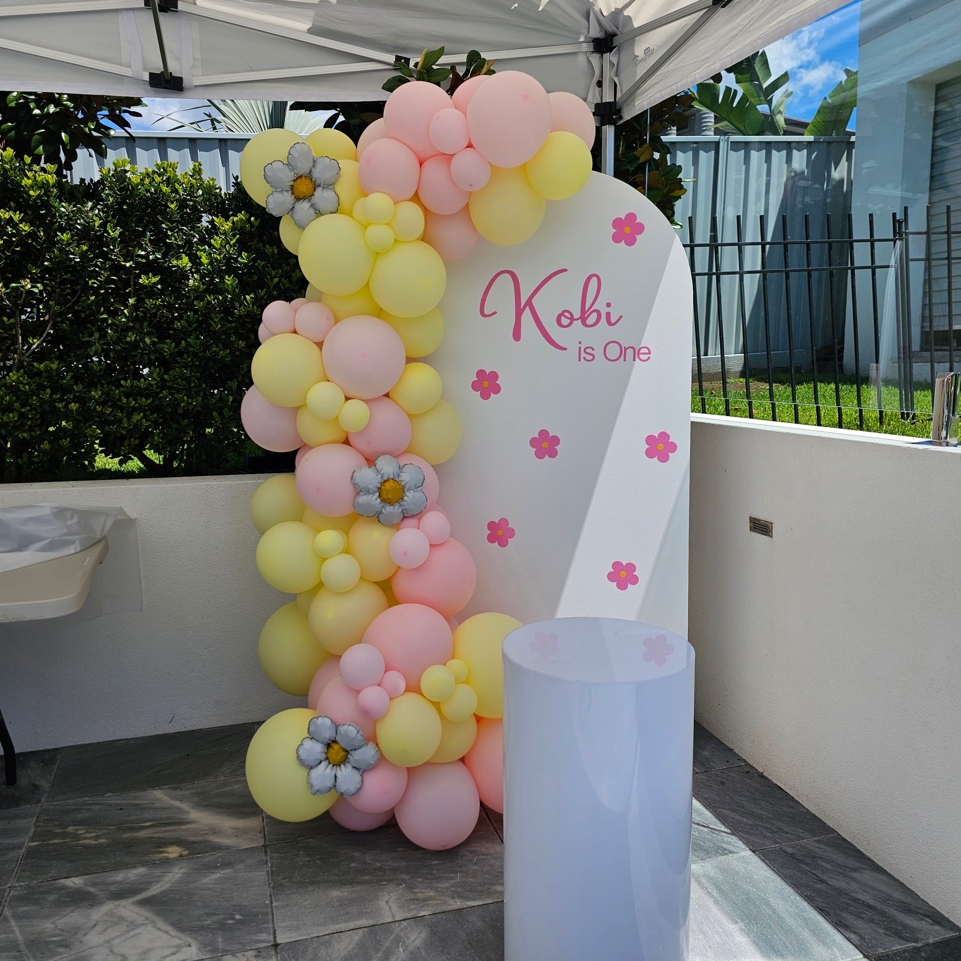 Daisy themed first birthday with white arch backdrop, with daisy flowers and personalisation, large white acrylic plinth and balloon garland decoration featuring daisy balloons