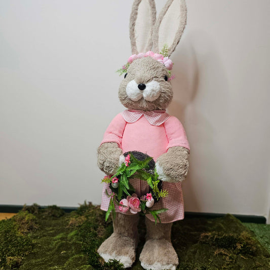 Stuffed bunny prop in a pink outfit with a flower wreath, standing on a green surface against a white wall.