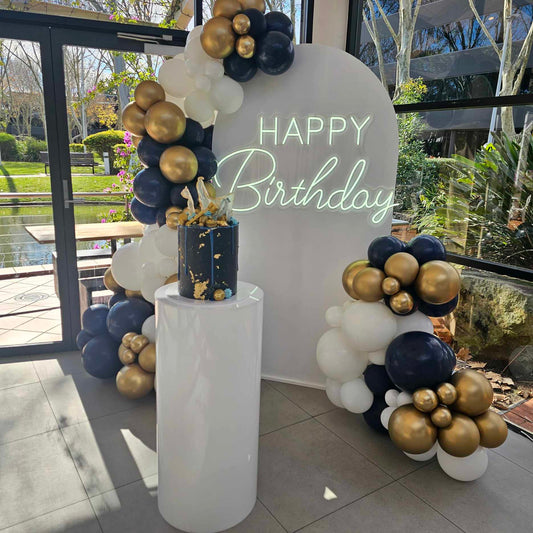 Happy Birthday Neon sign for hire, pictured with our white arch backdrop and White acrylic plinth and organic balloon garland.
