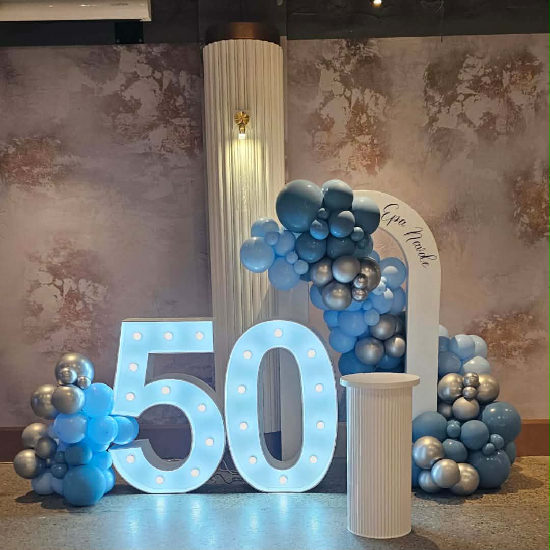 Decorative setup with blue and silver balloon garland, a large light up number '50', large ripple plinth and an Open arch backdrop against a textured wall.