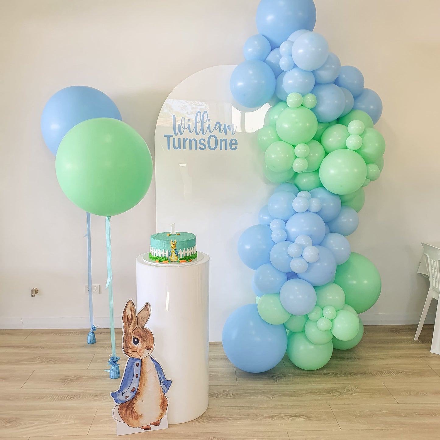 Peter Rabbit themed first birthday party featuring our personalised white arch backdrop, peter rabbit cutout and white acrylic plinth for the birthday cake. Jumbo Balloons in pastel mint and green with matching balloon garland decoration.